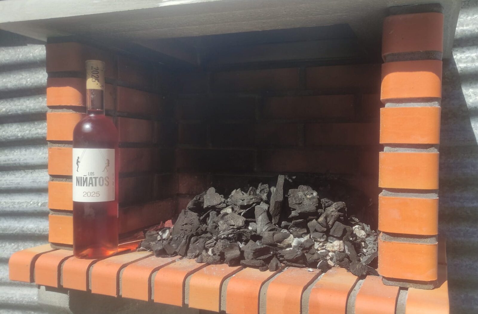Una botella del vino clarete Los Niñatos 2025 delante de una barbacoa en preparación
